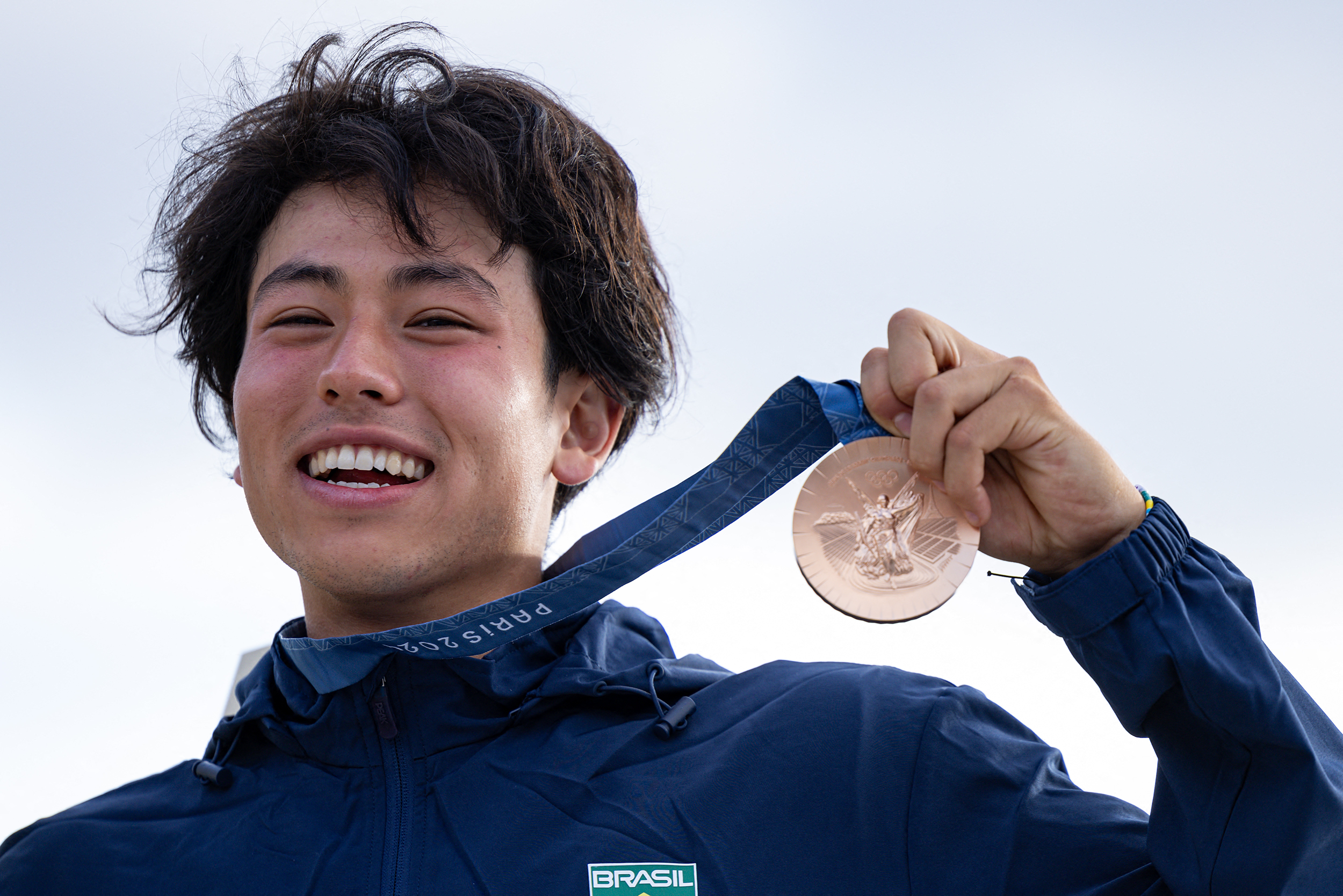 Fotografia de busto de um homem de etnia asiática sorrindo enquanto segura uma medalha de bronze. Ele usa blusa na cor azul.