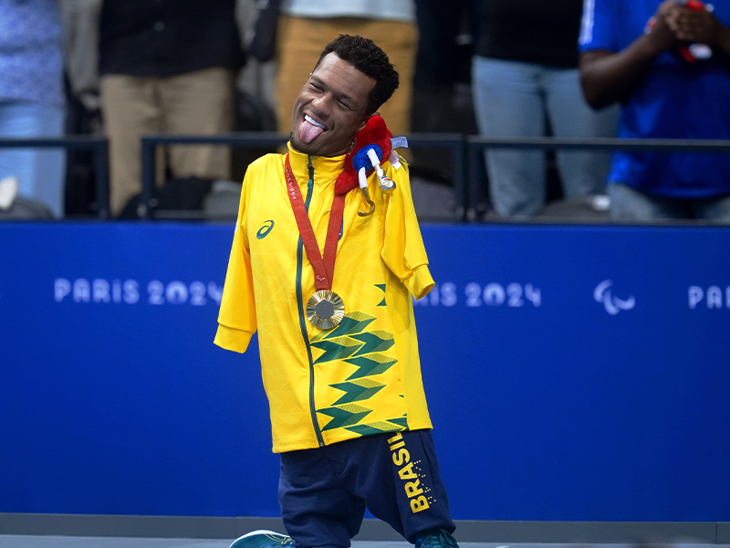 Fotografia de um homem sorrindo com a língua para fora. Ele não tem os braços, está com uma medalha no pescoço e usa blusa amarela com detalhes verdes e calça azul com a palavra Brasil escrita em amarelo. Entre o ombro e o pescoço, está segurando a mascote das paraolimpíadas. Ao fundo, há várias pessoas em uma arquibancada.