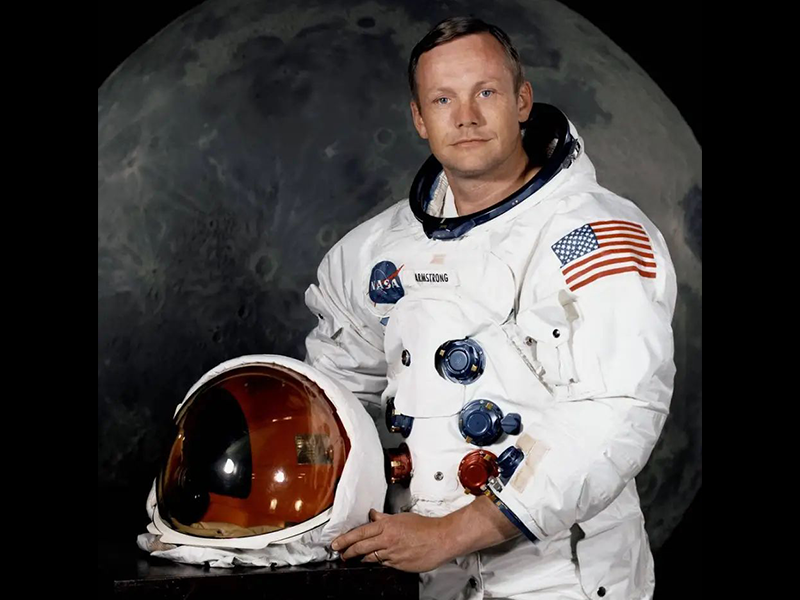 Fotografia de um homem visto da cintura para cima, olhando para a frente enquanto está posando para a foto. Ele usa roupa branca, de astronauta, e segura um capacete de astronauta. Sua roupa tem a bandeira dos Estados Unidos no ombro esquerdo e o logotipo da Nasa à direita, na frente. Ao fundo, a foto da Lua.