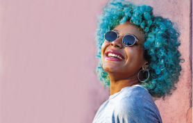 Fotografia de uma mulher sorrindo próxima a uma parede rosa. Ela tem cabelos volumosos e cacheados em cor azul, usa óculos de sol redondos com lentes escuras, brincos grandes de argola e usa camiseta clara.