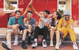 Fotografia de cinco jovens sentados lado a lado e todos sorrindo. Da esquerda para direita, está um jovem de cabelos curtos e castanhos, usando camisa, shorts e tênis; uma jovem com cabelos bem curtos, com jaqueta jeans e botas pretas; um jovem de cabelos pretos, usando óculos, regata e tênis, e com uma bola de basquete nos pés. Ao lado dele está uma jovem de cabelos compridos, roupas coloridas e tênis. E por último está uma jovem com tranças longas e loiras, usando um chapéu, roupas amarelas e tênis.