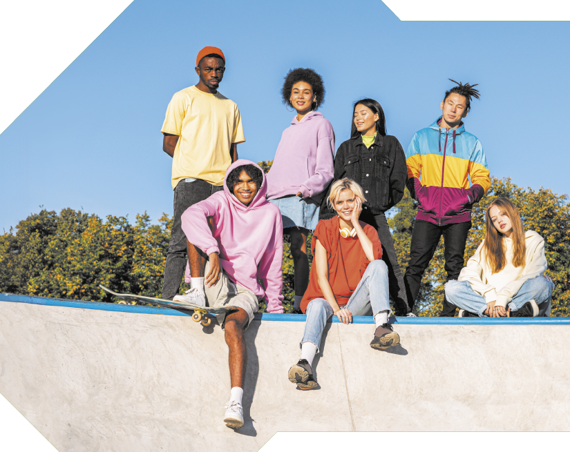 Fotografia de sete jovens no alto de uma pista de skate. À frente, sentados, está um rapaz com moletom rosa e shorts e com um pé apoiado em um skate. Ao lado está uma jovem com cabelos curtos e loiros, com fones de ouvido no pescoço e usando camiseta laranja e calça. Ao lado dela, está outra jovem, que tem cabelos compridos, usa moletom claro e calça. Atrás deles estão os outros quatro jovens de pé. Da esquerda para direita: um jovem com gorro e camiseta amarela; uma jovem com cabelos pretos, cacheados e volumosos usando moletom rosa; uma jovem com cabelos pretos e lisos, usando jaqueta jeans; e um jovem com tranças no cabelo em um peteado arrepiado, usando jaqueta colorida em azul, amarelo e rosa.