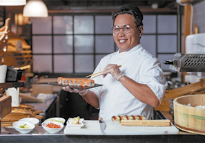 Fotografia de um homem em uma cozinha. Ele usa óculos, roupas brancas e luvas e está segurando hashis em uma mão e um prato de sushi na outra. À frente dele há uma bancada com vários potes com outros alimentos.