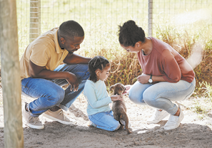 Fotografia de uma menina acariciando um pequeno cachorro marrom. Ela está agachada e entre um homem e uma mulher que estão agachados ao lado dela, olhando para o cachorro. Eles estão em um local com chão arenoso e com vegetação ao fundo cercado por telas.