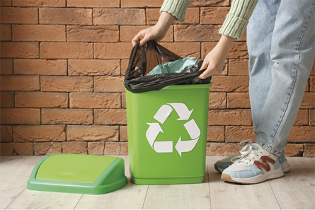 Fotografia de uma pessoa removendo um saco de lixo de uma lixeira verde que exibe o símbolo de reciclagem, composto por três setas formando um triângulo.