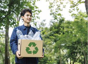 Fotografia de um jovem com cabelos castanhos, usando jaqueta azul. Ele está sorrindo enquanto segura uma caixa de papelão com o símbolo de reciclável, representado por três setas que formam um triângulo. A caixa está cheia de garrafas PET vazias. Ao fundo, há várias árvores.