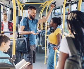Fotografia de um homem e uma mulher conversando em pé dentro de um ônibus. O homem está usando com roupas casuais, usando tem cabelos pretos, barba e bigode, usa camiseta, shorts e está com uma bolsa de alça lateral. A mulher está com camiseta amarela, calça jeans e fones de ouvido pendurados no pescoço. Ambos se seguram nos apoios do ônibus. Ao redor, algumas pessoas estão sentadas nos bancos e outras em pé.