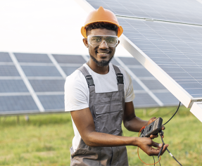 Fotografia de um homem com capacete e óculos de proteção, usando uma camiseta branca e um macacão. Ele segura um aparelho nas mãos e está próximo de placas solares.