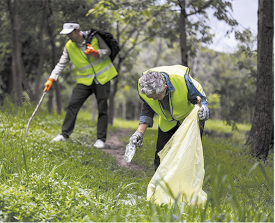 Fotografia de duas pessoas limpando uma área arborizada, durante o dia. Ambas estão usando coletes de segurança verde e luvas. Uma delas está curvada, segurando um saco de lixo amarelo e recolhendo uma garrafa plástica do chão. A outra está com uma mochila nas costas e segura um bastão de coleta. O ambiente ao redor é composto por árvores e grama.
