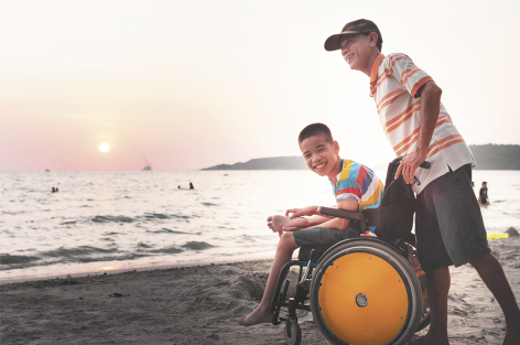 Fotografia de um menino e um homem em uma praia durante o pôr do sol. O menino está sentado em uma cadeira de rodas, sorrindo, enquanto o homem, está em pé atrás dele. Ao fundo está o mar e algumas pessoas pela praia.