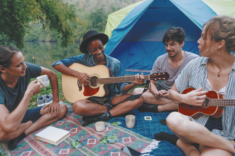 Fotografia de quatro pessoas sentadas perto de uma barraca azul e verde. Duas delas tocam instrumentos musicais, uma toca violão e a outra toca um instrumento semelhante, mas de tamanho menor. As outras duas pessoas observam e uma delas segura uma xícara. Eles estão sobre um pano estendido no chão, onde há um livro e copos. Ao fundo há várias árvores.