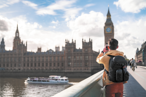 Fotografia de uma pessoa vistas de costas, com uma mochila fotografando a construção com a torre do relógio Big Ben, em Londres. Ela está sobre uma ponte, e ao lado, no rio há um barco navegando.