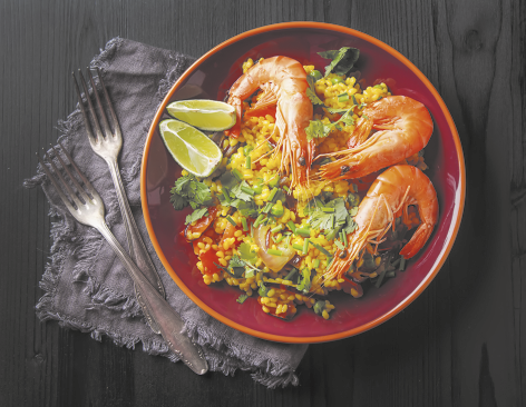 Fotografia de um prato com comida sobre uma mesa ao lado de dois garfos. Nele há arroz amarelo, pimentão e ervilhas, três camarões grandes e inteiros, com casca e cauda, folhas verdes e duas fatias de limão.
