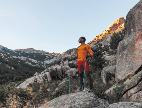 Fotografia de um homem em um ambiente montanhoso, no topo de uma rocha. Ele usa uma blusa laranja, shorts vermelhos, meias longas e botas de caminhada. Também carrega uma mochila e segura dois bastões. Ao fundo, há montanhas com formações rochosas e vegetação esparsa.