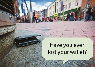 Fotografia de uma carteira preta caída em uma calçada de uma rua movimentada. Ao fundo, há várias pessoas andando, prédios e lojas. Sobre a foto há um balão de fala que diz em inglês: Have you ever lost your wallet?
