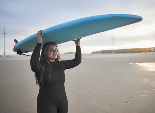Fotografia de uma mulher de cabelos claros e longos, usando roupas pretas e segurando uma prancha azul acima da cabeça. Ela está em uma praia de areia durante o dia.