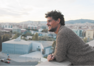 Fotografia de um homem de cabelos cacheados e barba curta, encostado em uma mureta de uma sacada, sorrindo enquanto observa a paisagem urbana. Ele usa blusa marrom e ao fundo há prédios, árvores e montanhas.