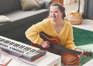 Fotografia de uma jovem com Síndrome de Down sorrindo enquanto toca violão. Ela está com cabelos presos, usa blusa amarela e está em uma sala com sofá ao fundo e um teclado a sua frente.