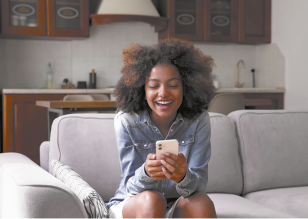 Fotografia de uma mulher com cabelo volumoso sentada em um sofá, segurando um celular. Ela está sorrindo enquanto olha para o celular, usa uma camisa jeans e ao fundo, há uma cozinha com armários de madeira.