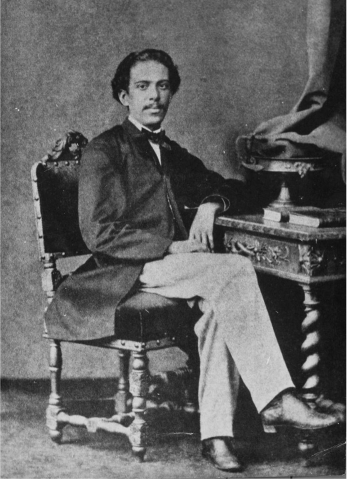 Fotografia em preto e branco de um homem sentado em uma cadeira, usando casaco escuro, gravata borboleta e calças claras. Ele está com o braço apoiado em uma mesa ornamentada ao seu lado.