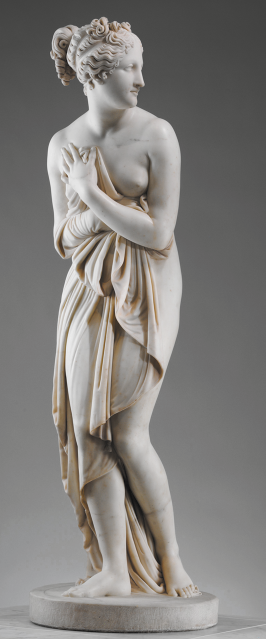 Fotografia de uma escultura representando uma mulher parcialmente coberta por um tecido. Ela tem o cabelo cacheado e preso, está com o rosto direcionado para à direita e com o corpo levemente curvado para frente, enquanto segura o tecido próximo ao peito, cobrindo um seio, e deixando o outro a mostra. O corpo da mulher é representado com cintura fina, quadris arredondados e pernas alongadas.
