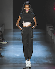 Fotografia de uma modelo desfilando em uma passarela. Ela é negra, tem cabelos pretos e longos e tem manchas ao redor da boca, na barriga e nos braços. Usa uma blusa preta curta, calça preta e sapatos brancos de salto.