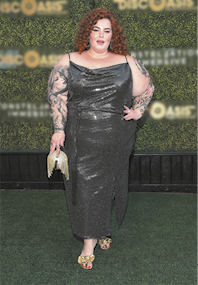 Fotografia de uma modelo plus-size. Ela é branca, tem cabelos ruivos e cacheados, tem braços tatuados, usa um vestido longo, escuro, brilhante e de alças finas. Está sobre um tapete verde, sorrindo com a mão na cintura e com um painel ao fundo.