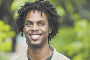 Fotografia de busto de um jovem sorrindo. Ele é negro, tem cabelos crespos e usa camisa clara.