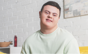 Fotografia de um jovem com Síndrome de Down sorrindo. Ele usa uma blusa clara e está em uma sala com um quadro na parede ao fundo.
