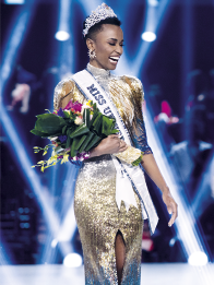 Fotografia de uma mulher sorridente, usando uma coroa brilhante e uma faixa que diz: MISS UNIVERSE. Ela é negra, usa brincos grandes, um vestido longo, dourado e brilhante, e segura um buquê de flores coloridas nos braços.