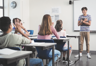 Fotografia de uma sala de aula com um grupo de estudantes. No centro, um jovem está em pé na frente da sala, com os braços cruzados e sorrindo. Vistos de costas, sentados em carteiras, estão outros alunos, os quais estão batendo palmas. Ao fundo há um quadro branco e alguns papéis na parede.