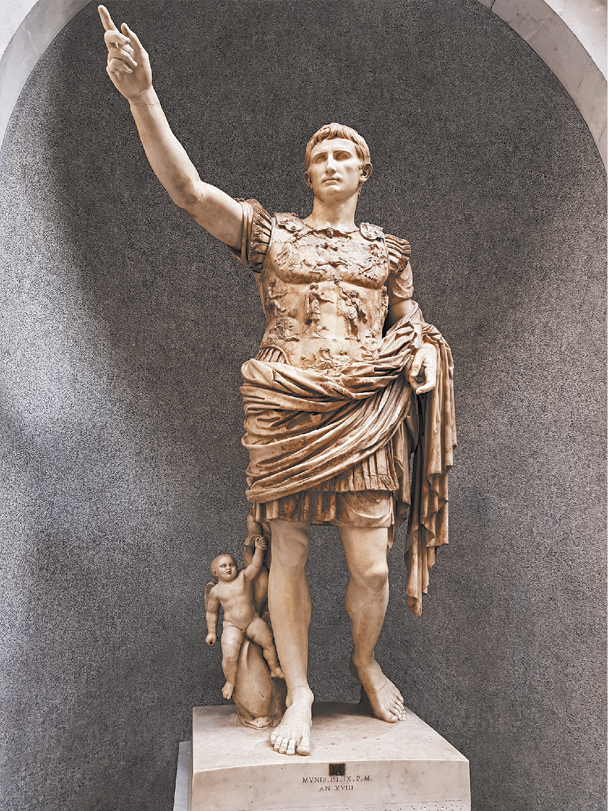 Fotografia de uma estátua de um homem, com o braço direito estendido e apontando para cima. Ele usa uma vestimenta detalhada com um peitoral contendo várias representações de figuras humanas, e usa um manto em torno do corpo. Ao lado do seu pé direito há uma pequena figura de um cupido montado em um golfinho.