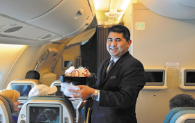 Fotografia de um homem em pé, sorrindo, no interior de um avião. Ele usa terno e gravata e segura uma bandeja com vários itens para os passageiros. Há várias poltronas, telas individuais e passageiros sentados.