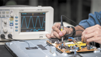 Fotografia de uma pessoa trabalhando em uma placa de circuito eletrônico. Ela está utilizando um equipamento semelhante a uma caneta sobre a placa e ao lado há uma tela exibindo um gráfico com várias curvas.