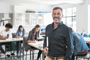 Fotografia de um homem em pé na frente de uma sala de aula. Ele está sorrindo, tem cabelos claros, barba grisalha e usa camisa preta. Atrás dele estão vários jovens sentados em carteiras.