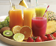Fotografia de uma bandeja repleta de frutas frescas e sucos coloridos. Há quatro copos de suco: um laranja, um amarelo, um verde e um vermelho. Ao redor dos copos, estão dispostas frutas como laranjas, morangos, kiwi, uvas, limão e pedaços de abacaxi.