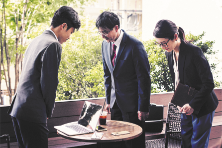 Fotografia de três pessoas em pé ao redor de uma mesa. São dois homens e uma mulher, e usam terno e blazer. Um dos homens está de frente para a mulher e o outro homem, e todos estão com o corpo curvado para frente e com a cabeça baixa. Sobre a mesa há um laptop aberto e um copo com bebida.