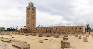 Fotografia de uma área ampla e externa com uma construção. No lado esquerdo, há uma torre alta e retangular e pontiaguda no topo. Ao lado, aparece uma estrutura longa e baixa composta por várias arcadas. À frente dessa construção há bases de colunas e estruturas de pedra.