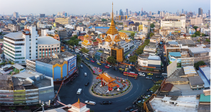 Fotografia com vista aérea de uma cidade com muitos edifícios. No centro, há uma rotatória com um monumento decorativo e ruas com veículos em movimento. Ao fundo, uma estrutura dourada em forma de templo se destaca em meio as outras construções.