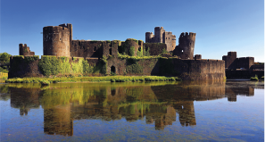 Fotografia de um castelo medieval com paredes de pedra cobertas em algumas partes por vegetação verde. O castelo está cercado por um corpo d’água que reflete sua estrutura, e o ambiente ao redor possui vegetação.