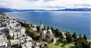 Fotografia com a vista aérea de uma cidade localizada às margens do mar, cercado por montanhas ao fundo. Em destaque, há uma catedral com uma alta torre central, situada em uma área arborizada. Ao redor, há muitos edifícios, construções e ruas.