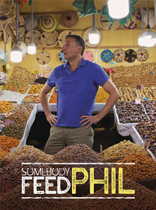 Capa de uma série intitulada em inglês: Somebody Feed Phil. No centro, há um homem de cabelo curto, usando uma camisa polo azul e calça clara. Ele está de pé, com as mãos na cintura, olhando para o lado, e ao seu redor há uma grande variedade de alimentos secos, como grãos e especiarias, e que estão dispostos em pilhas. Ao fundo, há decorações coloridas penduradas, como cestos de palha e outros enfeites artesanais. O homem está de pé, com as mãos na cintura, olhando para o lado com uma expressão pensativa.