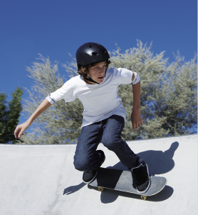 Fotografia de um menino andando de skate em uma pista. Ele está usando capacete camiseta branca, calça jeans e tênis. Seu corpo está inclinado para frente, e os dois pés estão sobre o skate. Ao fundo, há uma árvore.