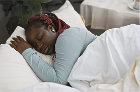 Fotografia de uma mulher dormindo em uma cama com travesseiros e coberta por um edredom. Ela tem tranças no cabelo nas cores vermelho e preto.