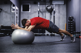 Fotografia de um homem em uma academia realizando um exercício utilizando uma bola de estabilidade. Ele está inclinado para frente, apoiando os antebraços na bola, enquanto mantém o corpo reto em uma posição de prancha. O fundo apresenta equipamentos de academia, como barras e halteres.