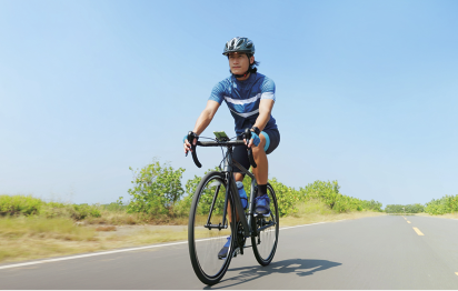 Fotografia de uma pessoa pedalando em uma bicicleta em uma estrada vazia e asfaltada, rodeada por vegetação sob um céu claro. Ela usa capacete, camiseta azul, bermuda de ciclista e luvas.