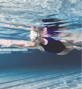 Fotografia de uma nadadora submersa em uma piscina, vista por dentro da água. Ela usa touca, óculos e um maiô escuro. Seu corpo está alinhado na água, com um braço estendido à frente e o outro para trás.