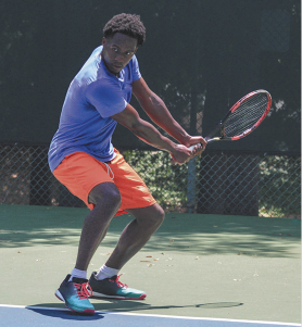 Fotografia de um homem jogando tênis em uma quadra ao ar livre. Ele usa camiseta azul, shorts laranja e tênis colorido, e está com os joelhos levemente flexionados, o corpo inclinado para frente e os dois braços segurando a raquete. Ao fundo, há uma cerca de malha metálica e árvores.