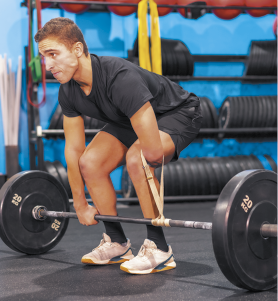 Fotografia de um homem em uma academia realizando um exercício de levantamento de peso. Ele está em posição de agachamento, segurando uma barra de peso no chão, com discos de 20 quilos em cada lado, usa camiseta preta, shorts pretos e tênis. Ele não tem uma das mãos e há um suporte elástico ligado aos seus antebraços e à barra. Ao fundo, há outros equipamentos, como anilhas empilhadas, barras e faixas elásticas.