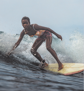 Fotografia de uma jovem surfando em uma onda. Ela está inclinada sobre a prancha, com os joelhos levemente dobrados, usa traje colorido e está descalça.